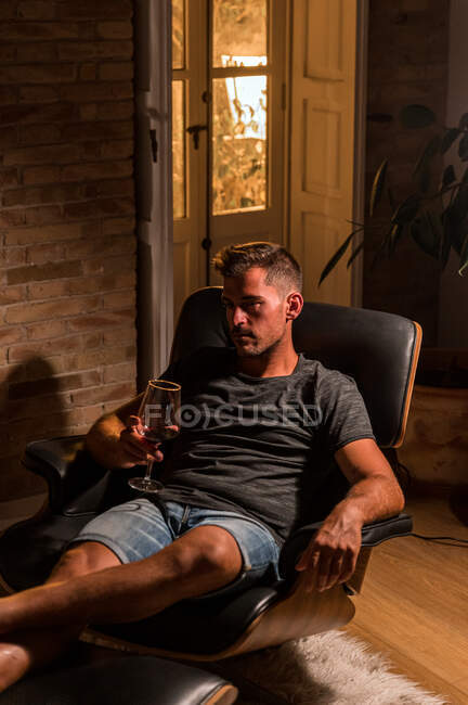 Vista lateral do macho em roupa doméstica refrigeração em poltrona confortável e beber vinho tinto enquanto desfruta da noite no quarto escuro — Fotografia de Stock