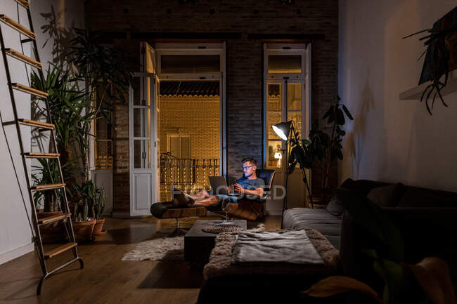 Umtriebiger männlicher Unternehmer sitzt im Sessel und arbeitet an einem Projekt, während er Laptop und Smartphone benutzt — Stockfoto