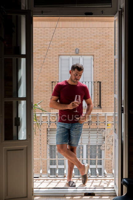 Розслаблений чоловік у домашньому вбранні стоїть на балконі і насолоджується свіжим білим вином — стокове фото