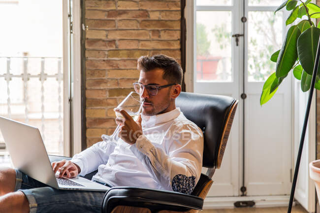 Beau entrepreneur masculin assis dans un fauteuil à la maison et travaillant sur un projet sur ordinateur portable tout en appréciant le vin — Photo de stock