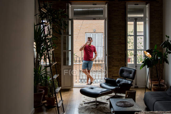Hombre relajado en traje doméstico de pie en el balcón y disfrutando de vino blanco fresco - foto de stock