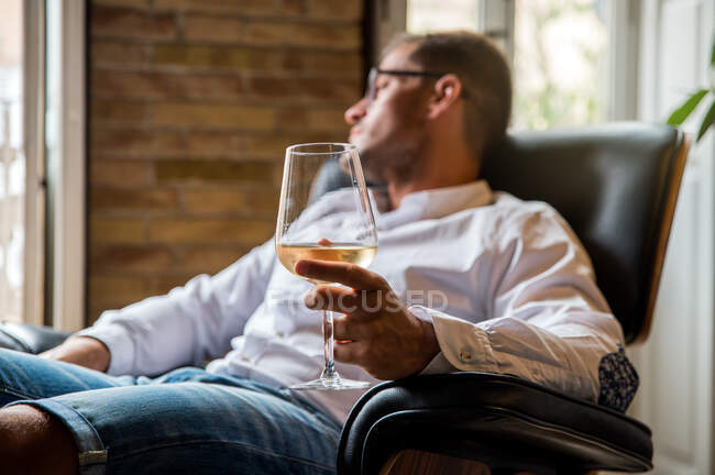 Богатый мужчина отдыхает в удобном кожаном кресле с бокалом белого вина и смотрит в раздумьях — стоковое фото