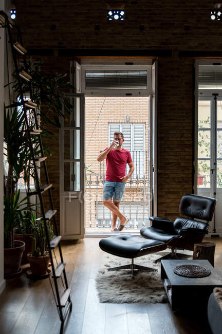 Расслабленный мужчина в домашней одежде стоит на балконе и наслаждается свежим белым вином — стоковое фото