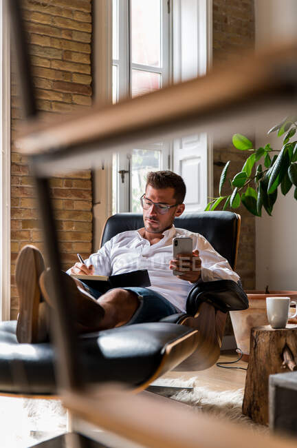 Imprenditore maschio concentrato seduto su una comoda poltrona e prendere appunti durante la pianificazione della giornata lavorativa a casa ufficio — Foto stock