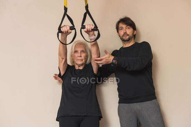 Adult male instructor behind senior female athlete during suspension training near sports equipment in gymnasium — Stock Photo