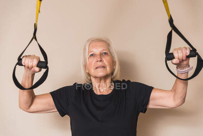Anciana atleta en ropa deportiva con canas haciendo ejercicio con correas mientras mira la cámara en el gimnasio - foto de stock
