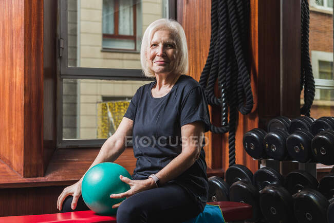Щаслива старша жінка-спортсменка з розумним годинником сидить на лавці з невеликим тренувальним м'ячем проти гантелів у гімназії, дивлячись на камеру — стокове фото