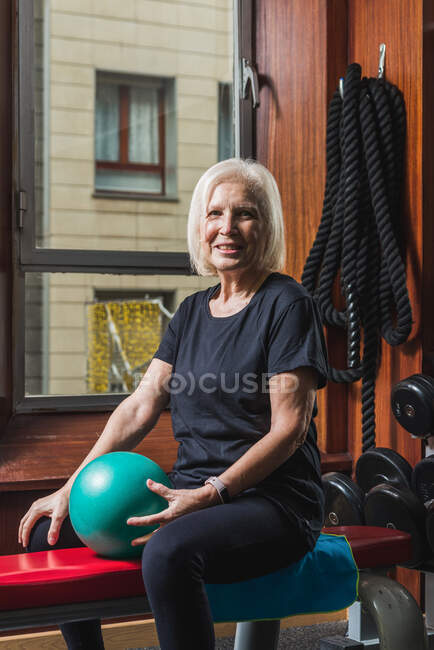 Glückliche Seniorin mit Smartwatch sitzt auf Bank mit kleinem Gymnastikball gegen Hanteln in Turnhalle und blickt in die Kamera — Stockfoto