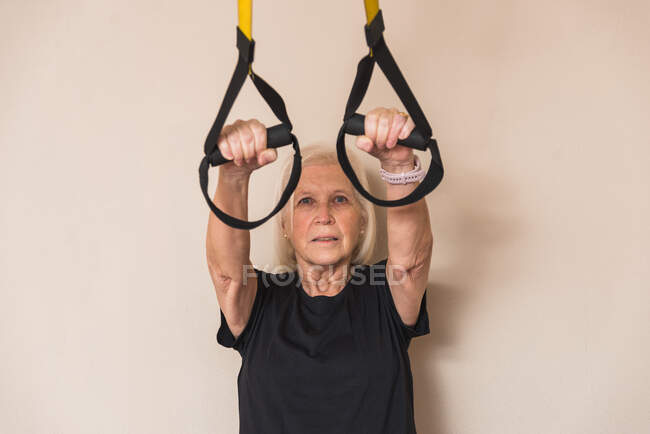 Elderly female athlete in sportswear with gray hair working out with straps while looking at camera in gym — Stock Photo