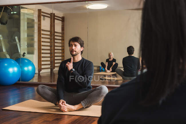 Adult barefoot male instructor with finger up explaining exercise to crop anonymous athletes while sitting on mat during Pilates training — Stock Photo