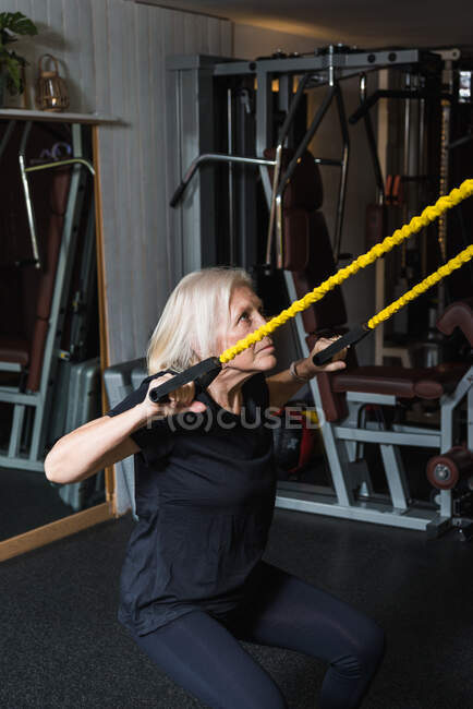 Vista laterale di anziana atleta donna in abbigliamento sportivo con capelli grigi che lavora con cinghie in palestra — Foto stock
