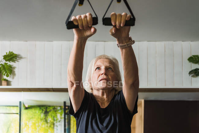 Focused elderly female athlete in sportswear with gray hair working out with straps while in the gym — Stock Photo