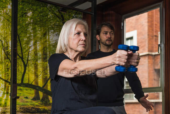 Atleta feminina determinada com cabelos grisalhos trabalhando com pesos perto do instrutor masculino enquanto olha para a frente no ginásio — Fotografia de Stock