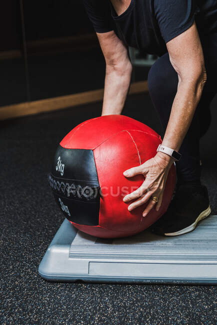 Crop anônimo atleta sênior feminino em roupas esportivas inclinando-se para a frente enquanto toma bola de remédio durante o treino no ginásio — Fotografia de Stock