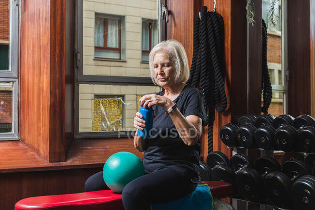 Garrafa de abertura de atleta feminina sênior enquanto sentada no banco com pequena bola de exercício contra halteres no ginásio — Fotografia de Stock