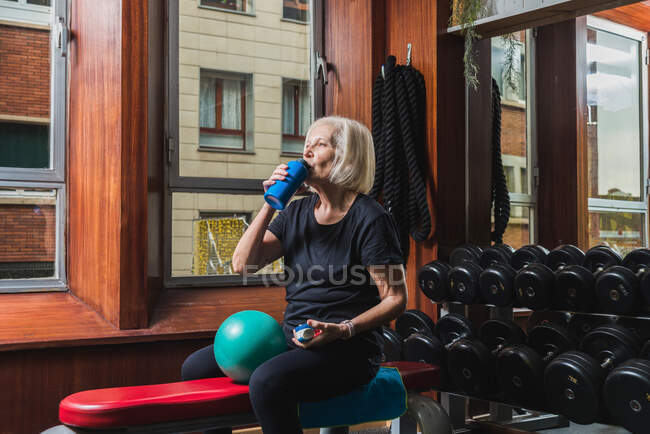 Durstige Seniorin trinkt Wasser aus Flasche, während sie mit kleinem Gymnastikball gegen Hanteln in Turnhalle sitzt — Stockfoto