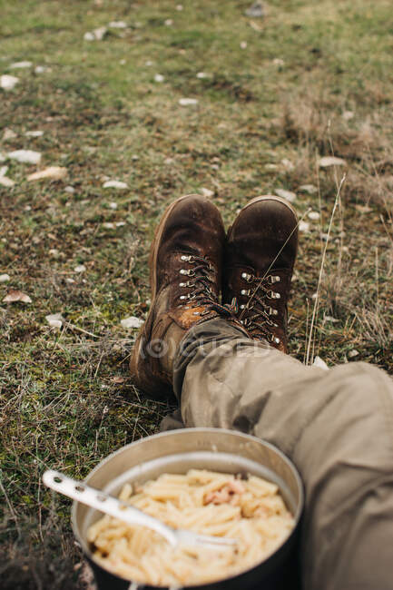 Обрізати анонімних чоловіків-мандрівників у чоботях, сидячи на землі з мискою макаронів під час подорожі на природі — стокове фото