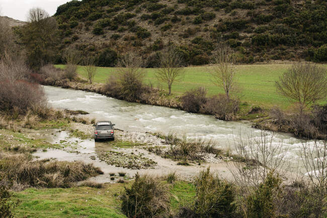 Сучасний позашляховик припаркований на березі швидкої річки, що тече в горбистій місцевості, покритій рослинами — стокове фото