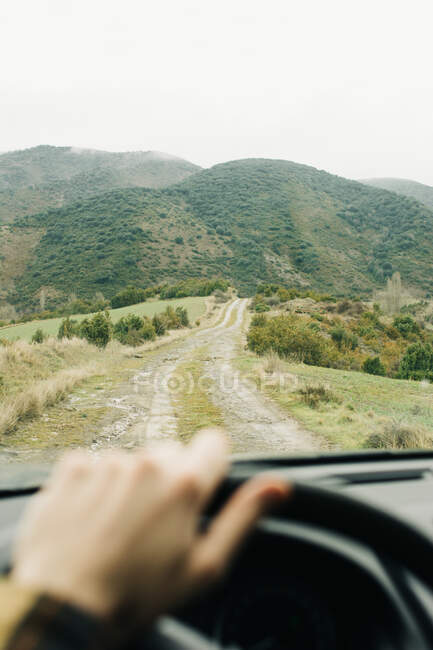 Crop anonymous male tourist driving car along empty rural road towards green hills during road trip — Stock Photo