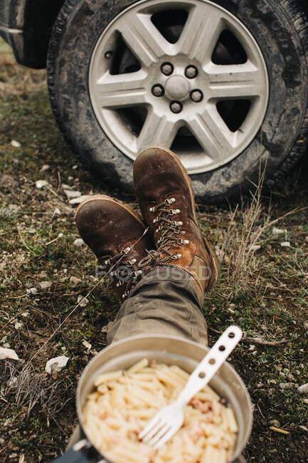 Crop anônimo viajante masculino em botas sentado no chão com tigela de macarrão durante a viagem na natureza — Fotografia de Stock