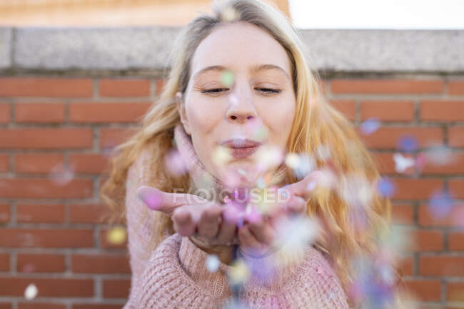 Heureuse jeune femme aux longs cheveux blonds soufflant des confettis colorés des mains tout en se tenant près du mur de briques le jour ensoleillé — Photo de stock