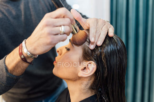 Cultive visagiste masculino irreconhecível usando escova enquanto aplica pó no rosto de modelo feminino étnico confiante — Fotografia de Stock
