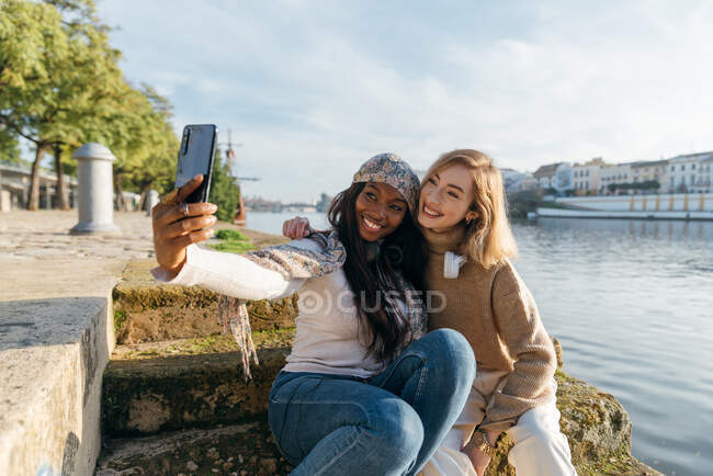 Content diverse female best friends sitting on stairs on promenade and taking selfie on smartphone during stroll on sunny day in city — Stock Photo