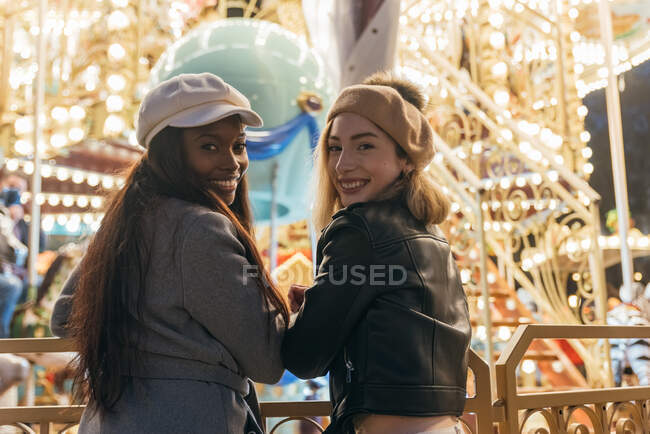 Rückansicht der entzückten multirassischen besten Freundinnen, die am Zaun des Karussells stehen, während sie das Wochenende im Freizeitpark verbringen und in die Kamera schauen — Stockfoto
