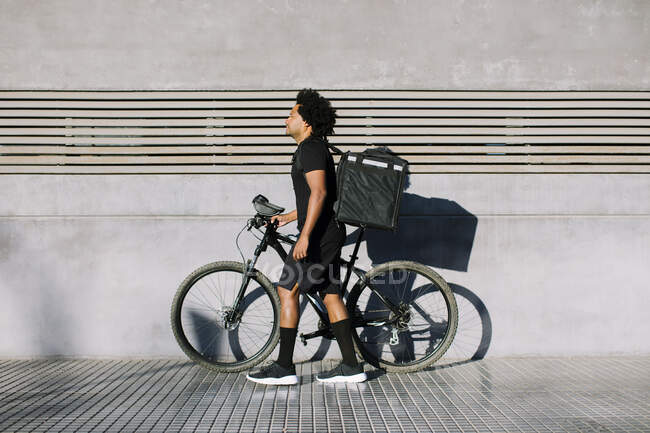 Vista laterale del cavaliere nero con scatola e bici che cammina sul marciapiede della città contro il muro con ombra mentre guarda lontano alla luce del sole — Foto stock
