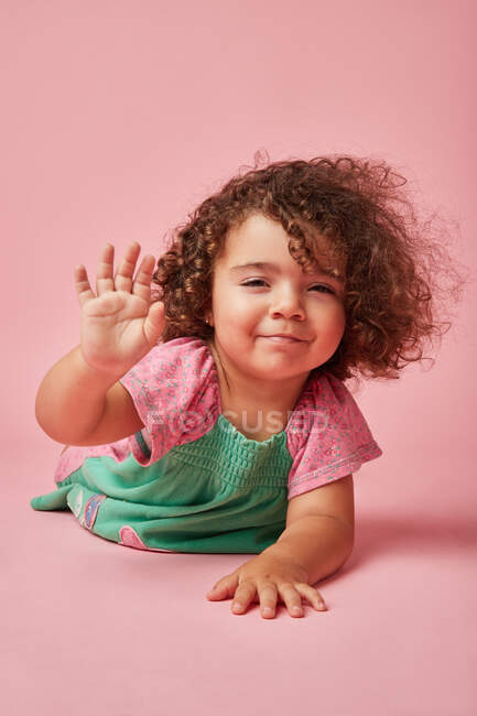 Adorabile bambino in abito con i capelli ricci guardando la fotocamera appoggiata con le mani sul pavimento che dice ciao con il gesto della mano — Foto stock