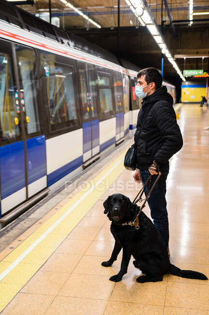 Вид сбоку под низким углом все тело анонимного слепого мужчины в маске стоящего с собакой-поводырем на поводке на платформе — стоковое фото