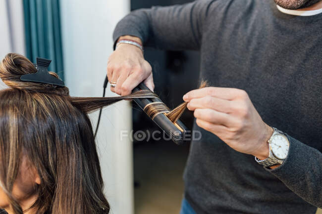 Cosecha estilista masculino irreconocible en ropa casual usando plancha de pelo mientras que hace rizos para cliente femenino en salón de belleza - foto de stock