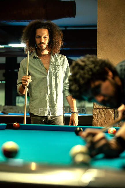 Serious ethic man with cue competing in game with player hitting billiard ball — Stock Photo