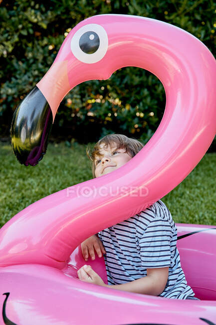 Веселый маленький мальчик в повседневной одежде сидит на надувном розовом фламинго, веселясь на травянистой лужайке в парке — стоковое фото