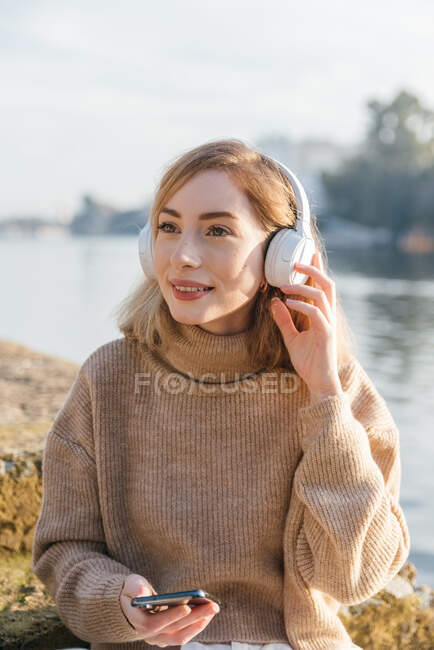 Mujer encantada sentada en el río y escuchando música en auriculares mientras disfruta de canciones - foto de stock