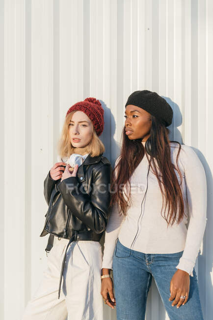 Cool multiracial female models wearing trendy clothes standing near metal wall in city on sunny day — Stock Photo