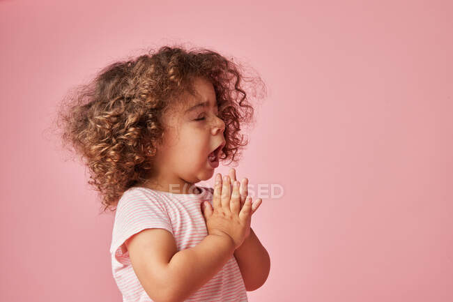 Vista laterale del bambino bambino carino con i capelli ricci starnuti con gli occhi chiusi e le mani in preghiera — Foto stock