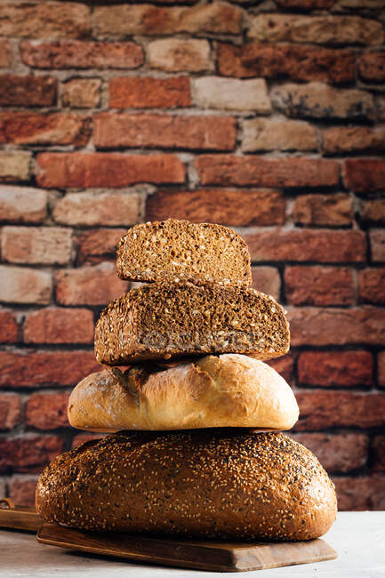 Pile di pane bianco e di segale con cereali e crosta appetitosa sul tagliere contro muro di mattoni in panetteria — Foto stock