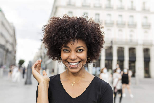 Joven mujer afroamericana alegre con peinado afro mirando a la cámara en la calle de la ciudad - foto de stock
