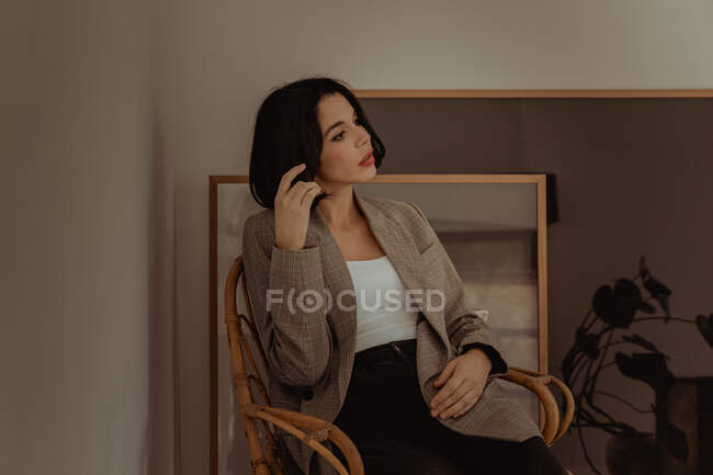 Удумлива жінка в модному одязі, сидячи на стільці, торкаючись волосся і дивлячись в роздуми — стокове фото