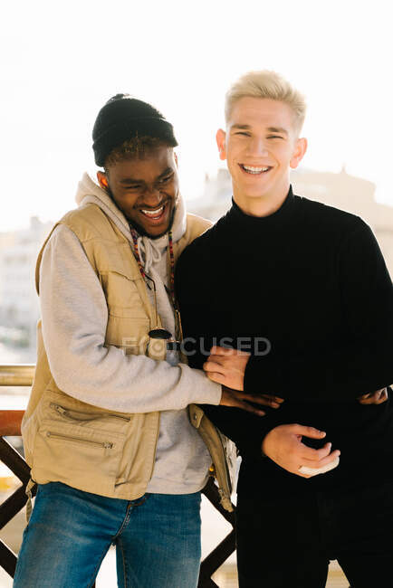 Joven alegre pareja gay multirracial en trajes de moda sonriendo y abrazándose mientras están juntos en la terraza en un día soleado - foto de stock