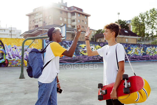Dos adolescentes con monopatín y mochila estrechando la mano y riendo en la calle - foto de stock