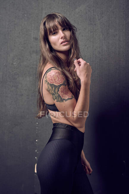 Чуттєва жінка з татуюванням у вигляді бюстгальтера, що торкається шиї в студії проти чорного фону. — стокове фото