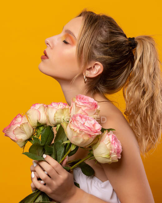Молодая неэмоциональная красивая женщина в белом платье с голыми плечами, держа нежные розовые розы, стоя с закрытыми глазами на желтом фоне в фотостудии — стоковое фото