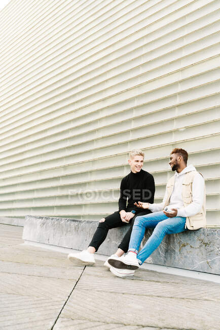 Full body of cheerful young multiethnic male friends in stylish clothes resting on bench in city park and chatting — Stock Photo
