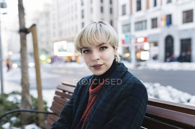 Jovem atraente com cabelo curto em casaco quente sentado no banco na rua contemporânea da cidade no final do dia de outono em Madrid, Espanha — Fotografia de Stock