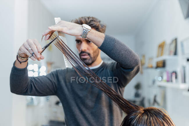Concentrated young ethnic bearded male master cutting hair of crop unrecognizable customer in modern salon — Stock Photo