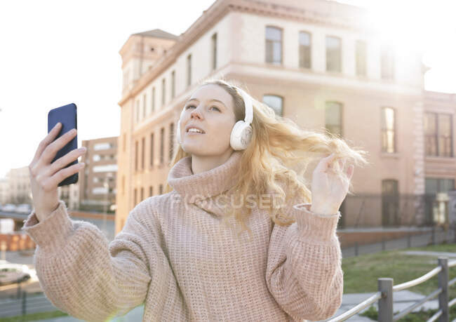 Selbstbewusste junge Frau in warmem Pullover und Kopfhörer berührt lange wellige Haare, während sie an sonnigen Tagen in der Stadt ein Selfie mit dem Smartphone macht — Stockfoto