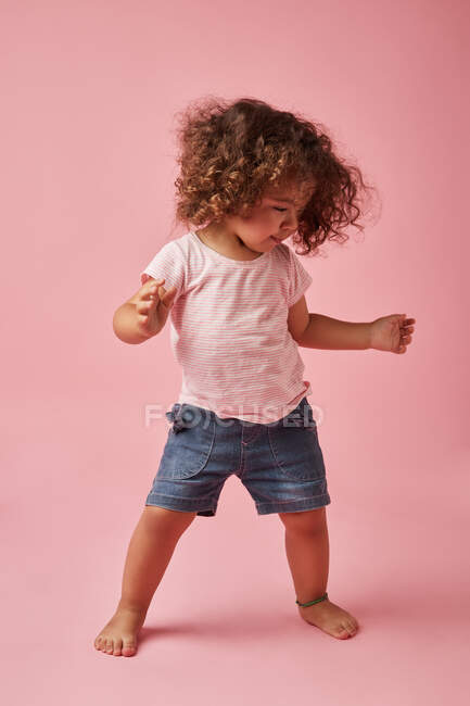 Очаровательный босоногий ребенок в футболке и джинсовых шортах с вьющимися волосами, смотрящий вниз во время танцев на розовом фоне — стоковое фото
