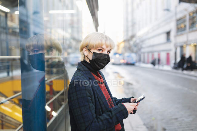 Спокійна молода жінка в теплому пальто і захисна маска, яка дивиться сучасний мобільний телефон, стоячи на вулиці міста в зимовий день в Мадриді, Іспанія. — стокове фото